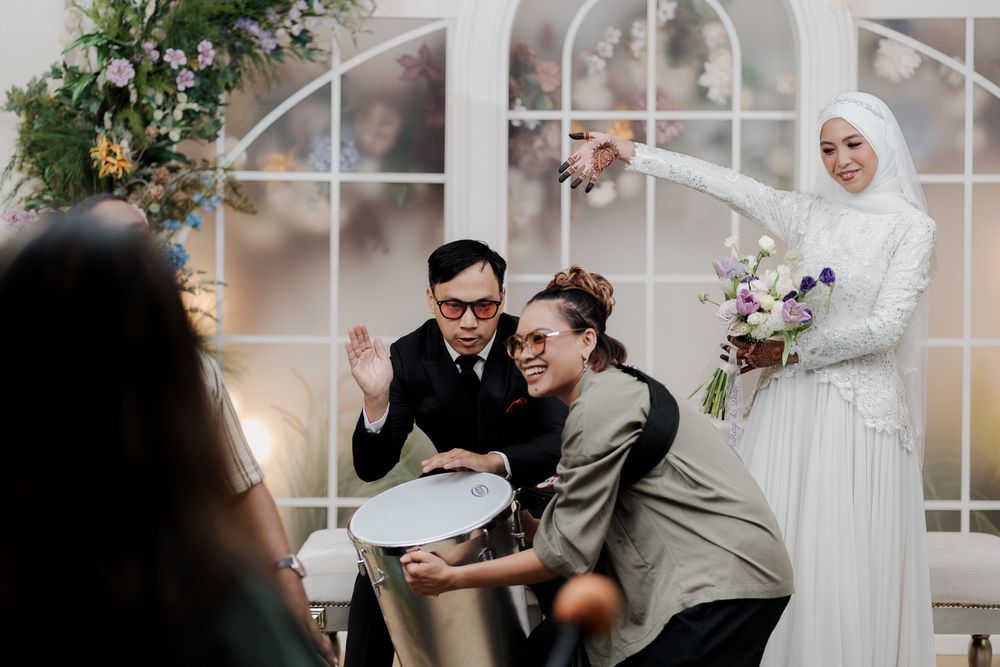 Wedding ceremony scene with guests laughing, bride in white hijab holding bouquet, elegant arch backdrop.