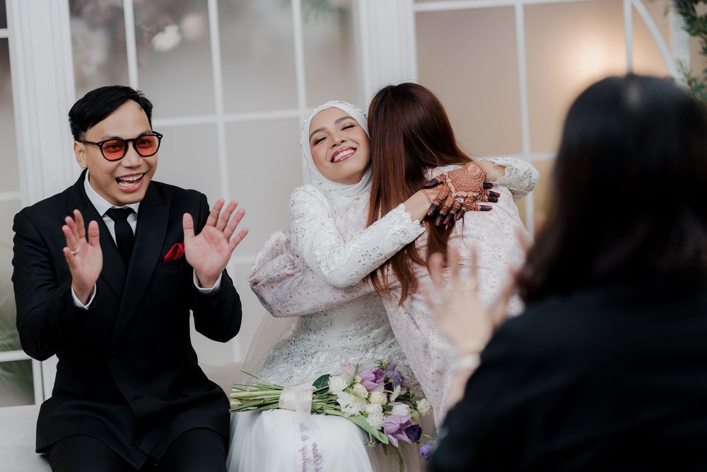 A joyful wedding scene with a bride in white hijab embracing a guest while a man in black suit claps happily.