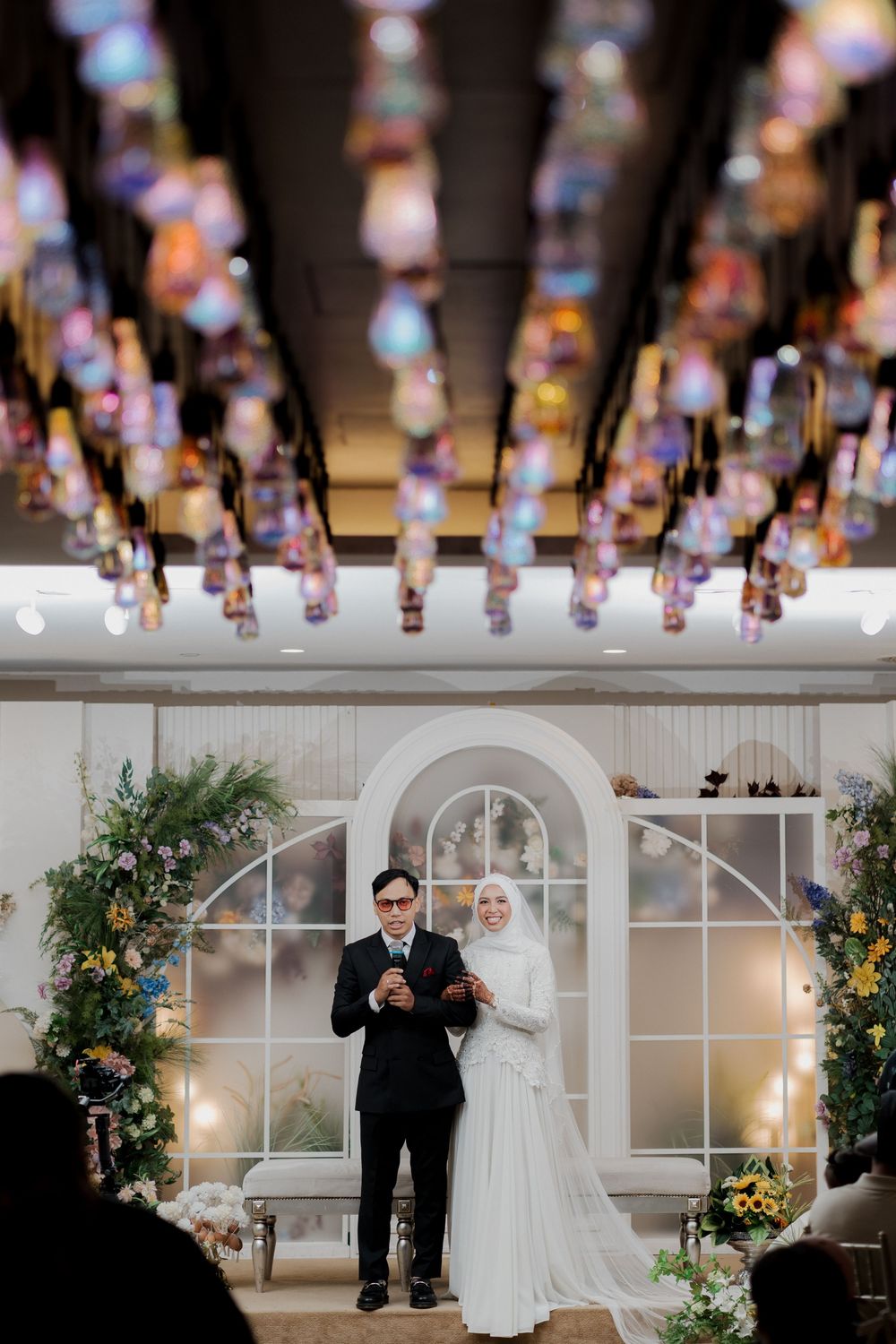 Elegant Muslim wedding ceremony with bride in white hijab gown and groom in black suit under colorful hanging lanterns.