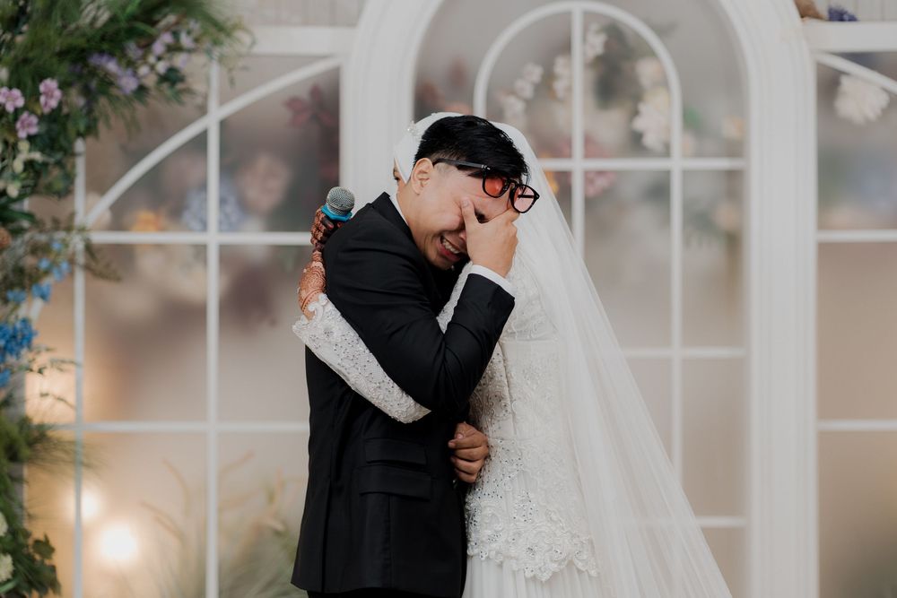 Emotional groom in black suit crying while embracing bride in white lace wedding dress and veil at altar.
