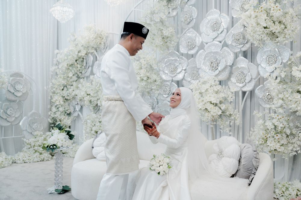 A couple in white traditional Malay wedding attire stands together surrounded by white floral decorations.