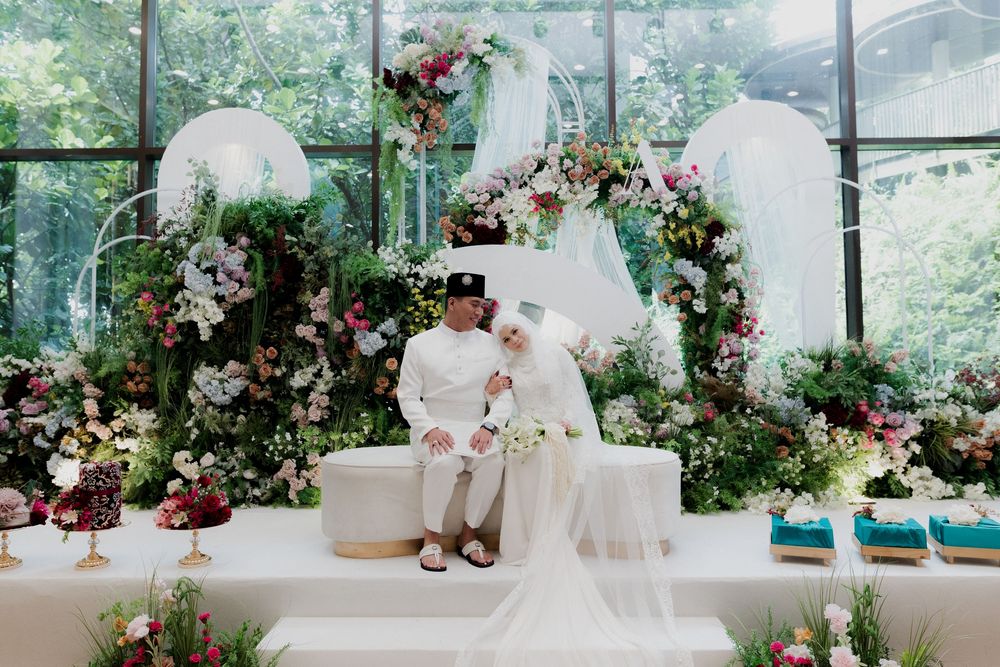 Muslim couple in white attire seated at a floral wedding reception backdrop with lush greenery and elegant flower arrangements.