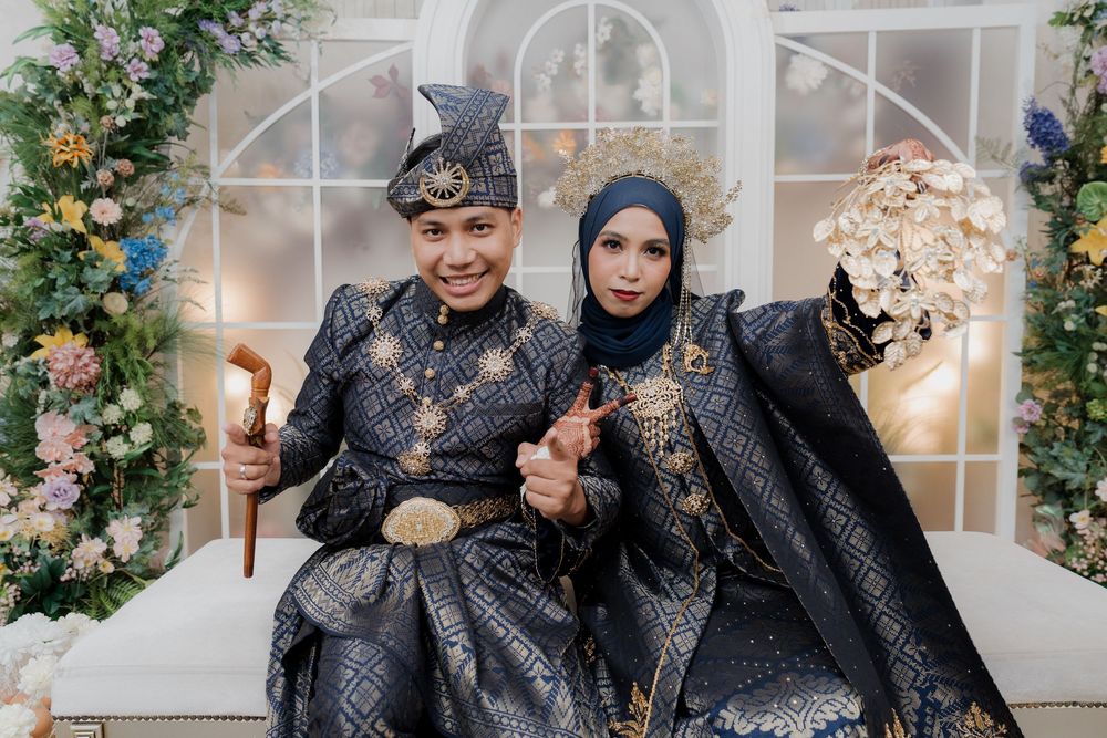 Couple in traditional Indonesian black and gold wedding attire with floral backdrop, posing together for a formal portrait.