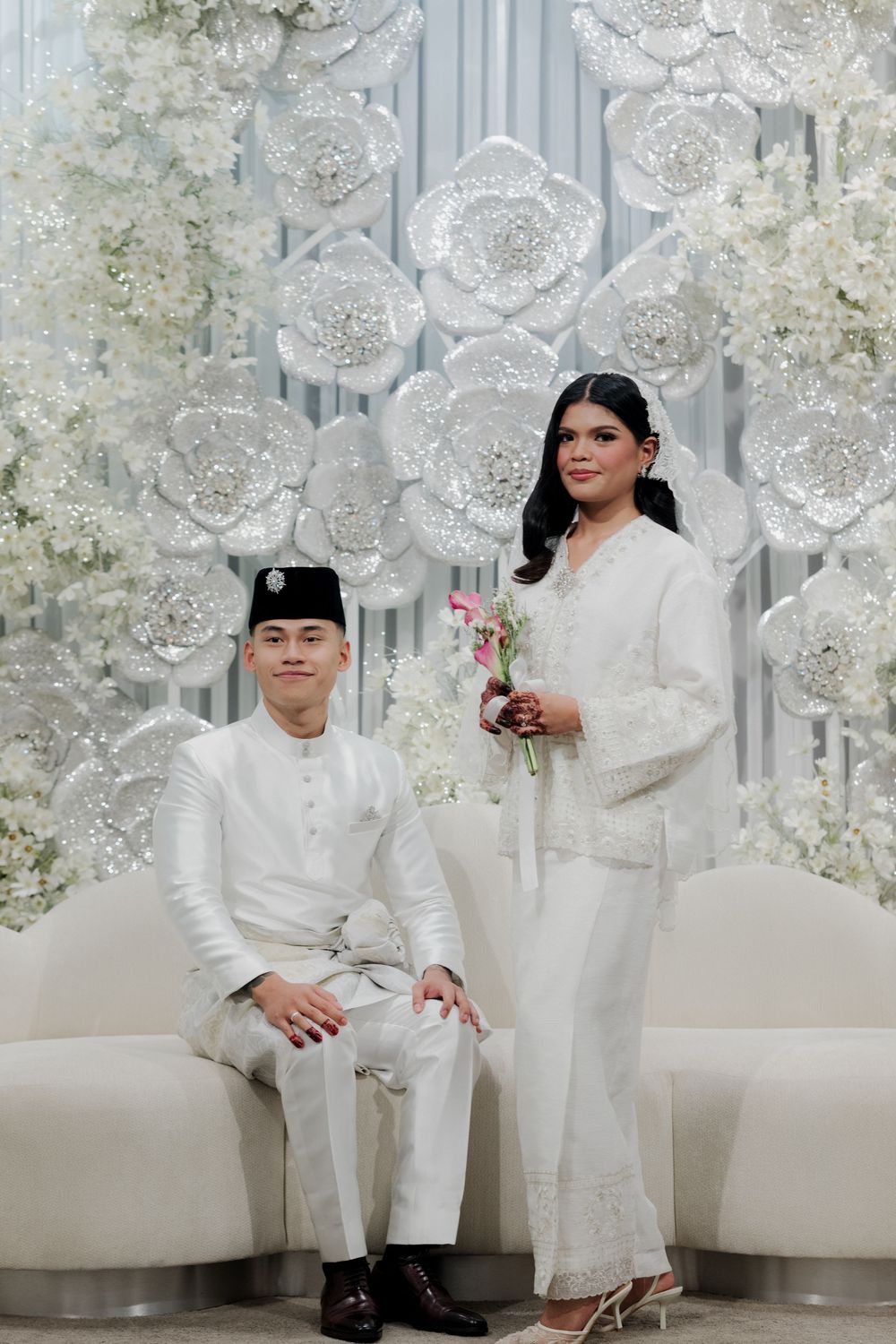 Malay couple in white traditional attire posing before elegant floral backdrop at a wedding ceremony.