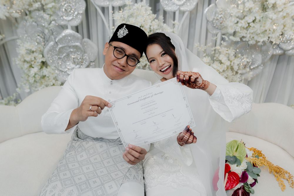 Couple in white traditional Malay attire holding marriage certificate at decorated wedding ceremony.