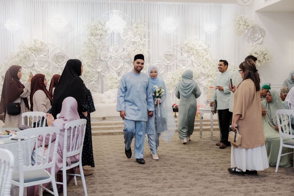 Muslim couple in traditional attire walks through elegant wedding reception hall with floral decor and guests.