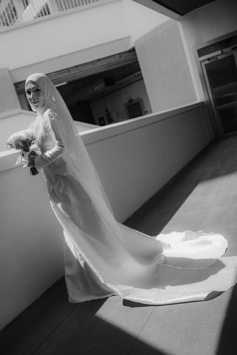 Black and white photo of a bride in a hijab and long white wedding dress with train, holding a bouquet.