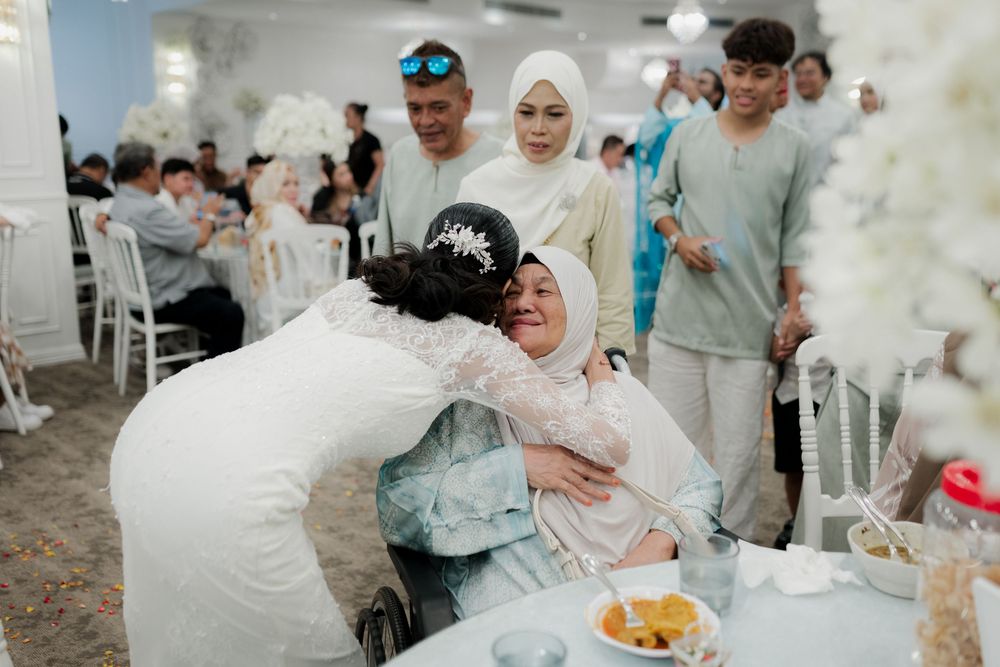 Bride in white hijab embraces emotional elderly woman in wheelchair at a Malay wedding reception.