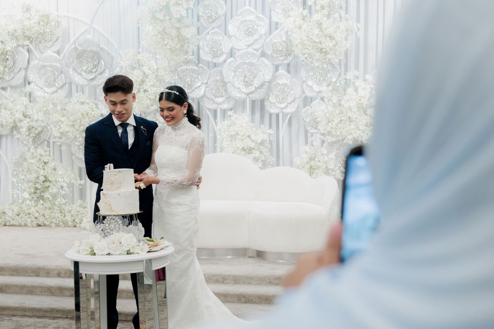 Bride and groom cutting wedding cake at elegant white-themed ceremony while guest captures moment on phone.