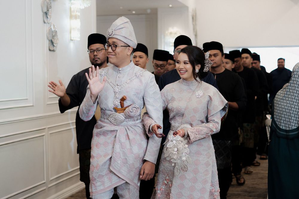 Malay couple in traditional wedding attire walking together at a formal ceremony with guests in background.
