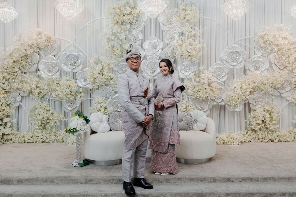 Couple in traditional Indonesian wedding attire posing before elegant white floral backdrop with roses and hydrangeas.