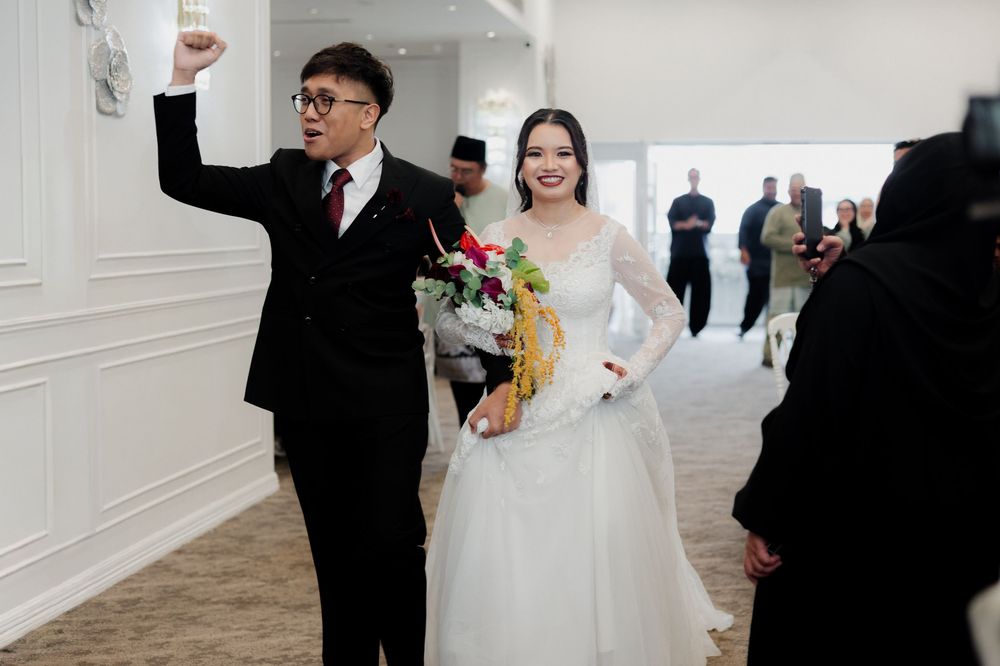 A bride in a white lace gown holds flowers beside a man in a black suit raising his fist at a wedding reception.