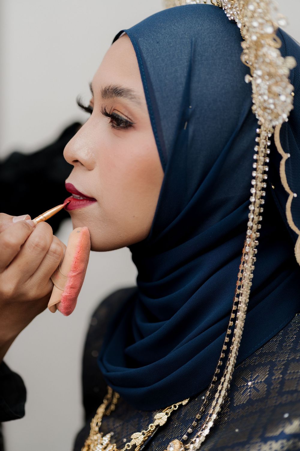 A Muslim bride in a navy hijab with gold embellishments gets red lipstick applied by a makeup artist.
