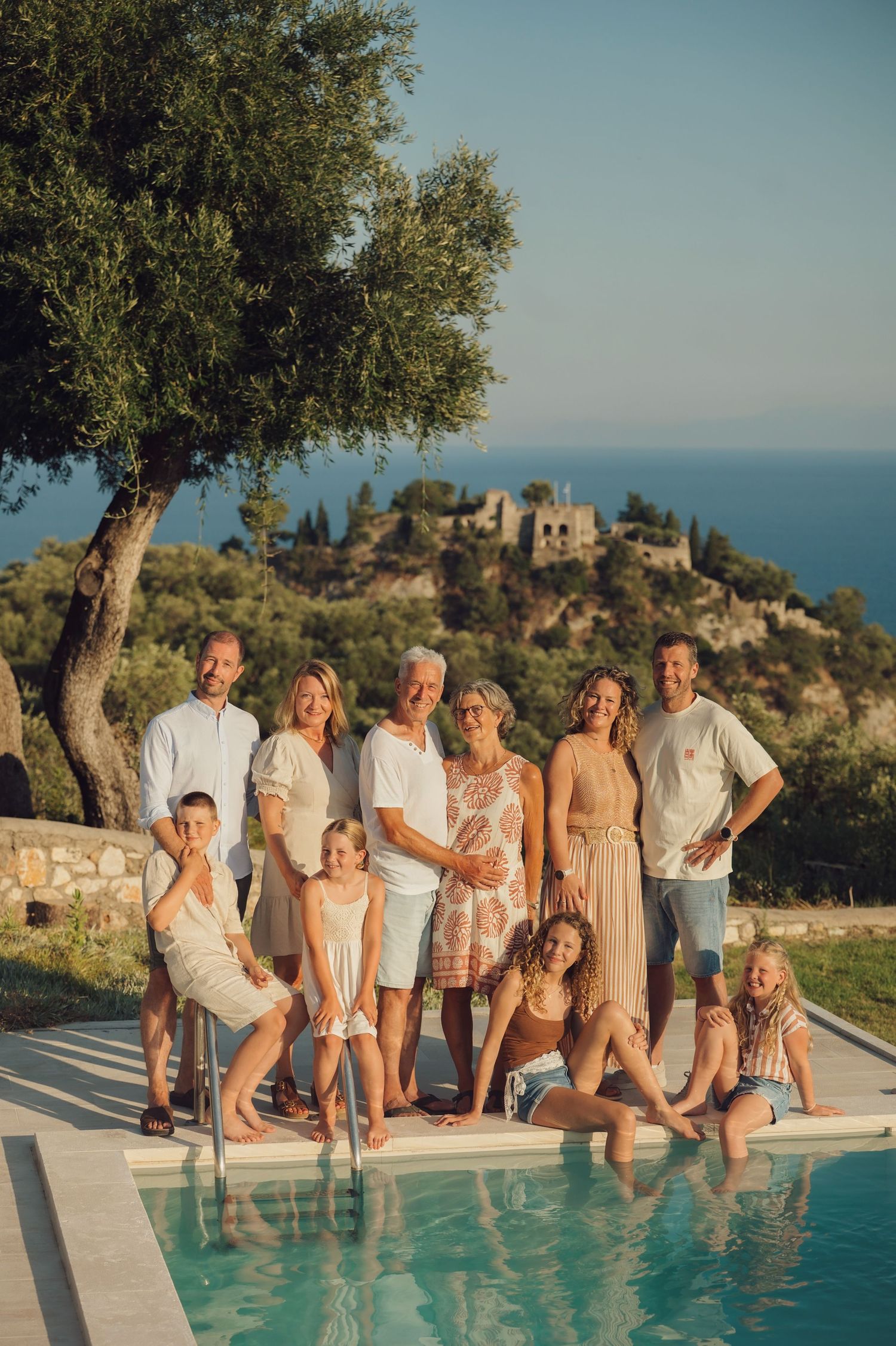 Multi-generational family portrait by a pool with Mediterranean sea views and lush greenery in warm golden light.