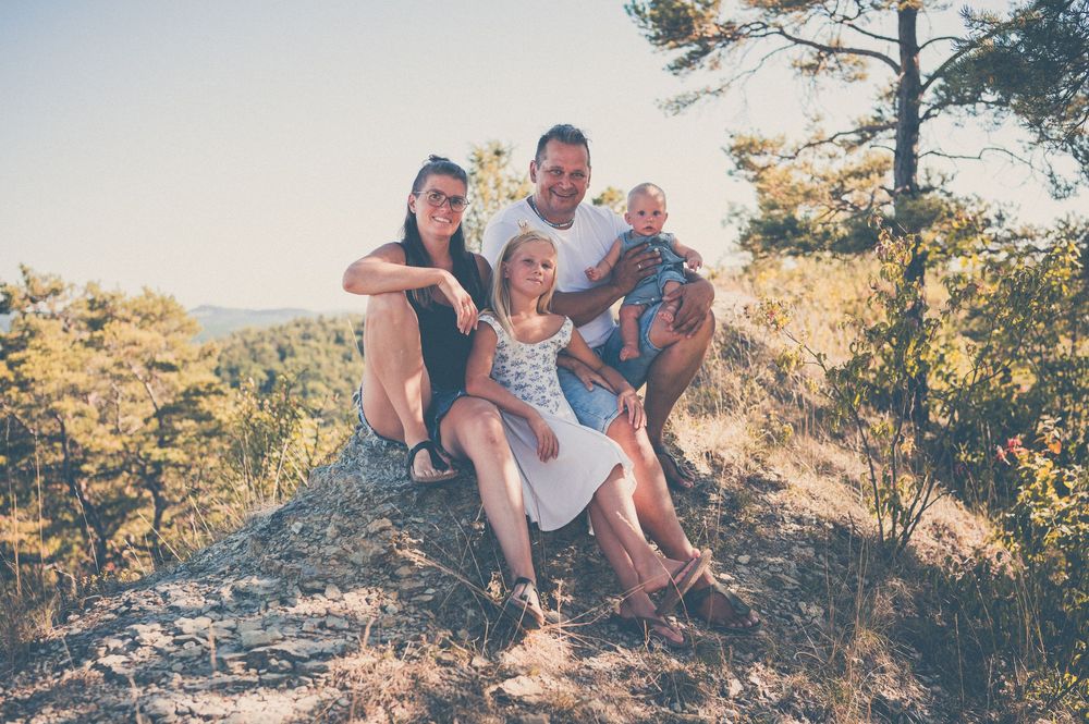 Family of four posing on a rocky hillside surrounded by lush green trees on a sunny day.