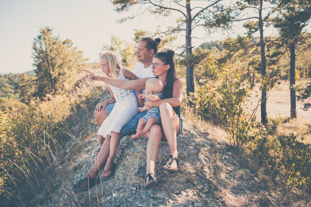 Family sitting together on a sunny hillside surrounded by trees, enjoying nature and pointing at the view.