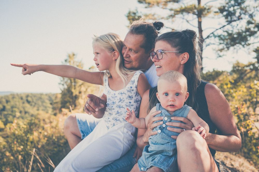 Happy family with two children sitting outdoors in nature, pointing at scenic view on a sunny day.