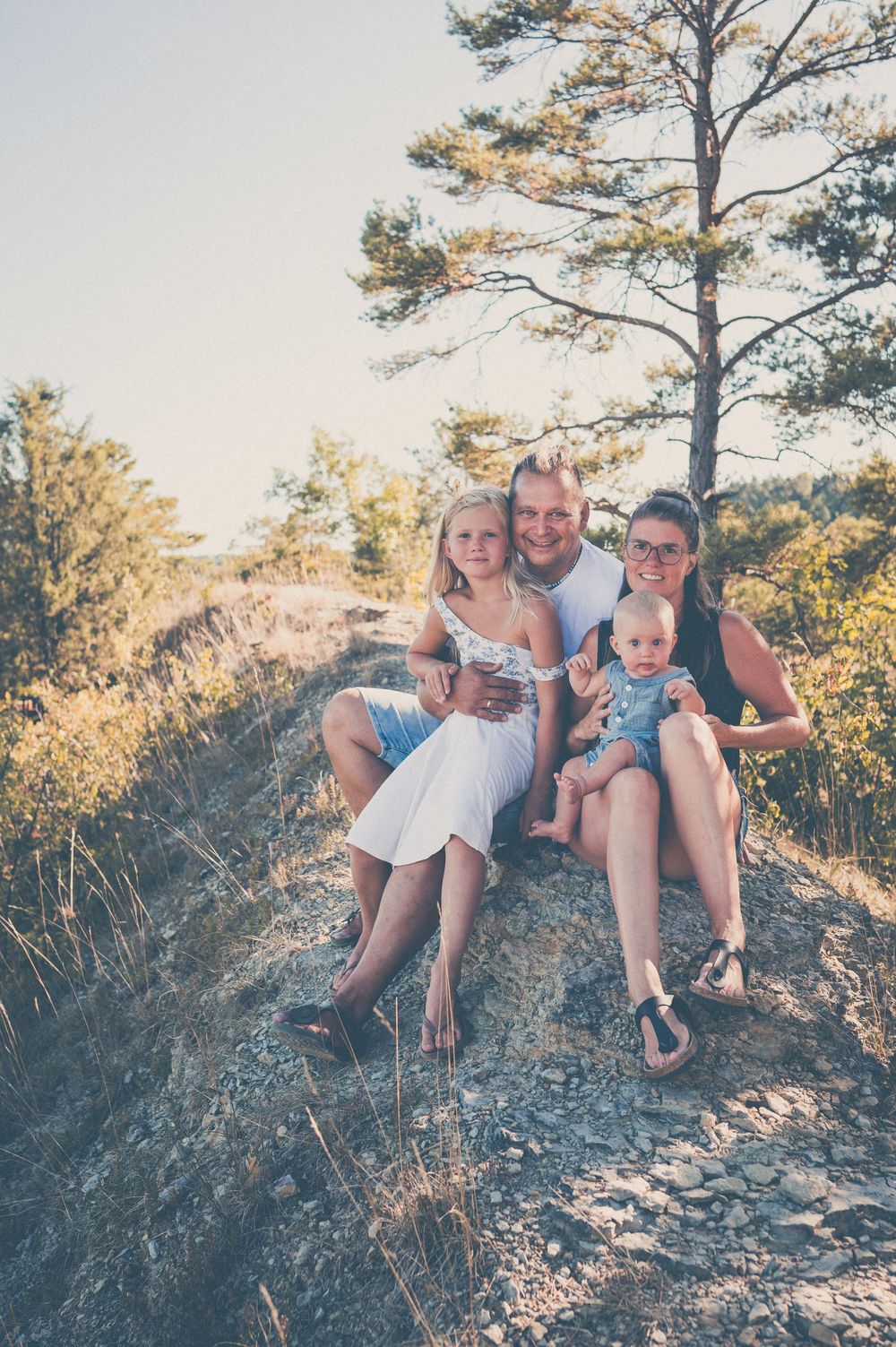 A family of four poses outdoors on rocky terrain near a pine tree on a sunny day.