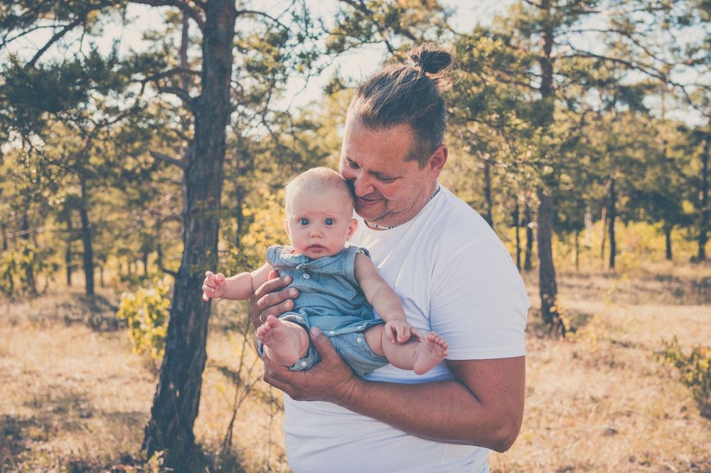 Father with man bun tenderly holding baby in blue outfit outdoors in sunny pine forest setting.
