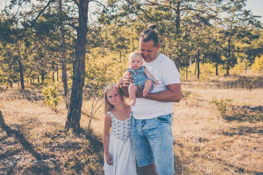A family of three poses outdoors in a sunny forest, with a man holding a baby and a young girl standing beside them.