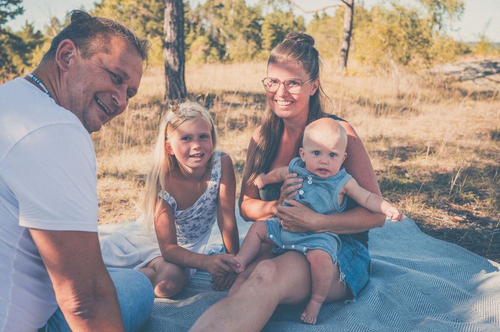 Family of four enjoying a sunny outdoor picnic, with two adults and two young children sitting on a blanket in a field.