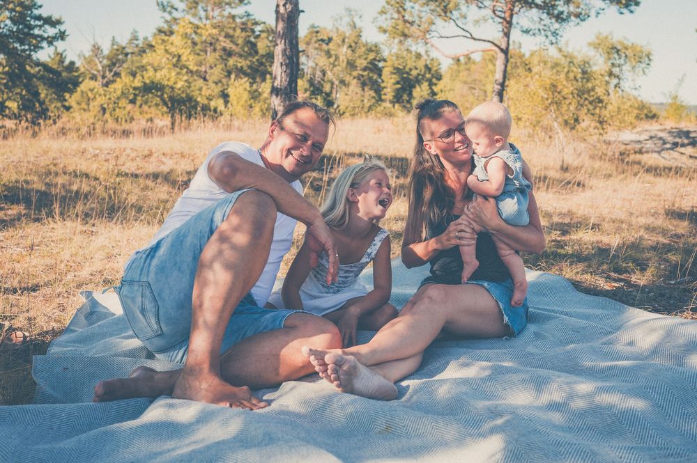 Family enjoying a sunny outdoor picnic on a blanket in a natural field setting with trees in the background.