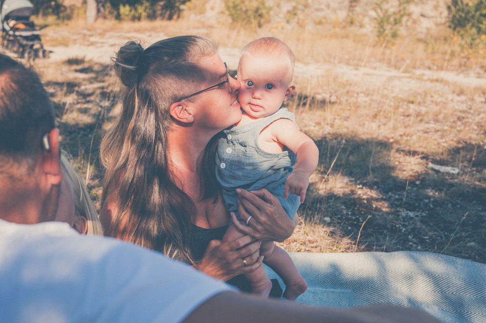 A mother tenderly kisses her baby outdoors in a sunny field during a family picnic.