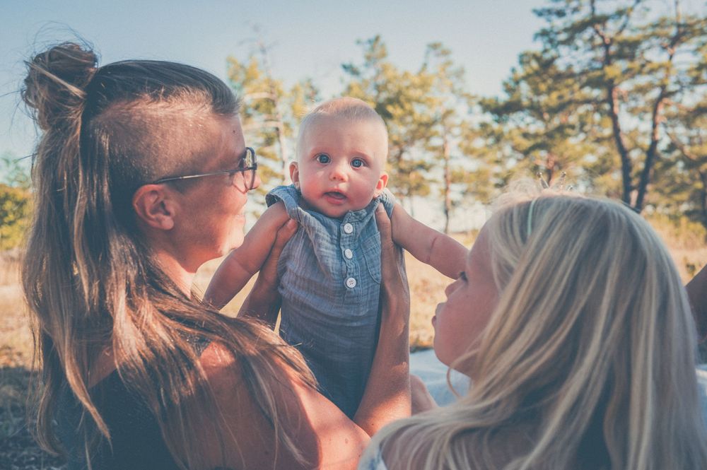 Two mothers lovingly hold their baby outdoors surrounded by pine trees on a sunny day.