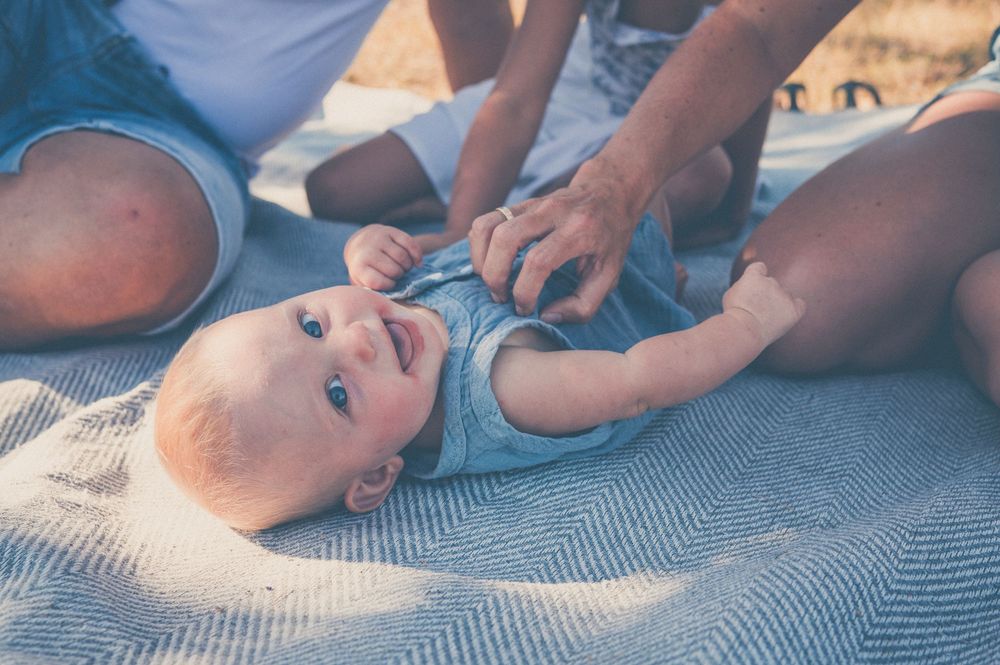Baby lying on blanket at beach being tickled by parents on sunny day.
