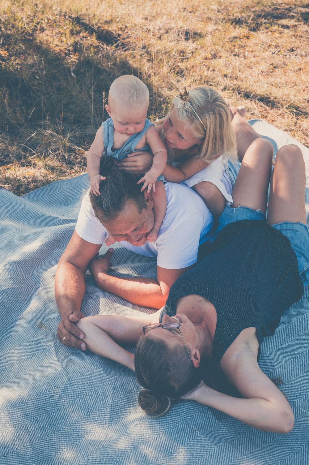 Family relaxing on a blanket outdoors, with two adults and two young children playing together on a sunny day.