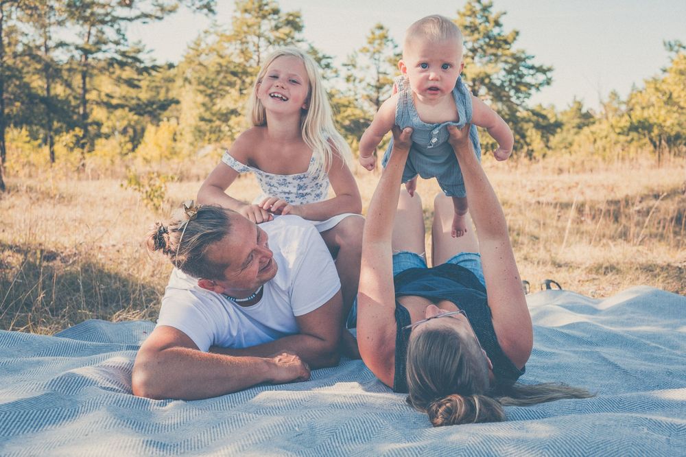 Family playing outdoors on a blanket in a sunny field, parents lifting laughing children in a joyful moment.