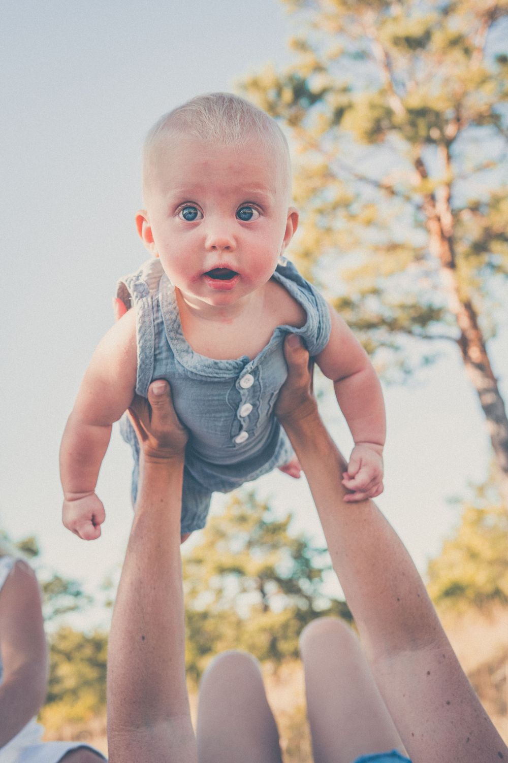 Adult hands lifting a wide-eyed baby in a blue romper outdoors with trees in the background.