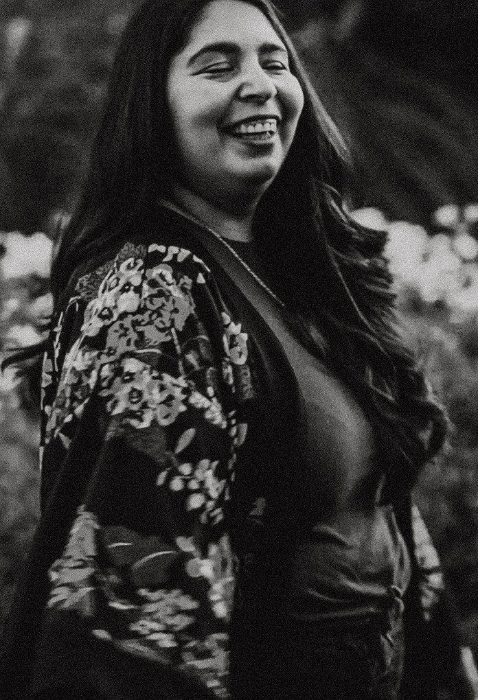 Black and white portrait of a smiling young woman with long hair wearing a floral jacket outdoors.