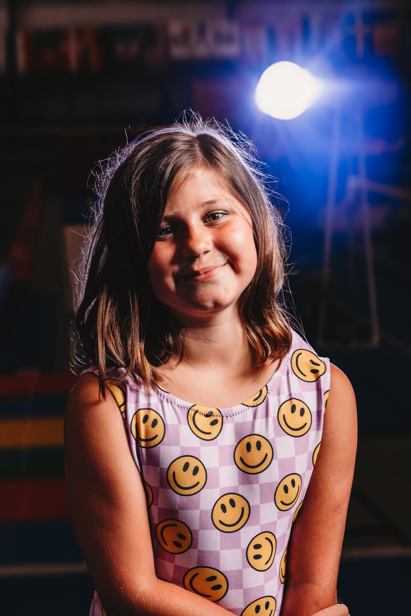 Young girl smiling in a smiley face patterned dress, posed under a bright stage light in a dark setting.