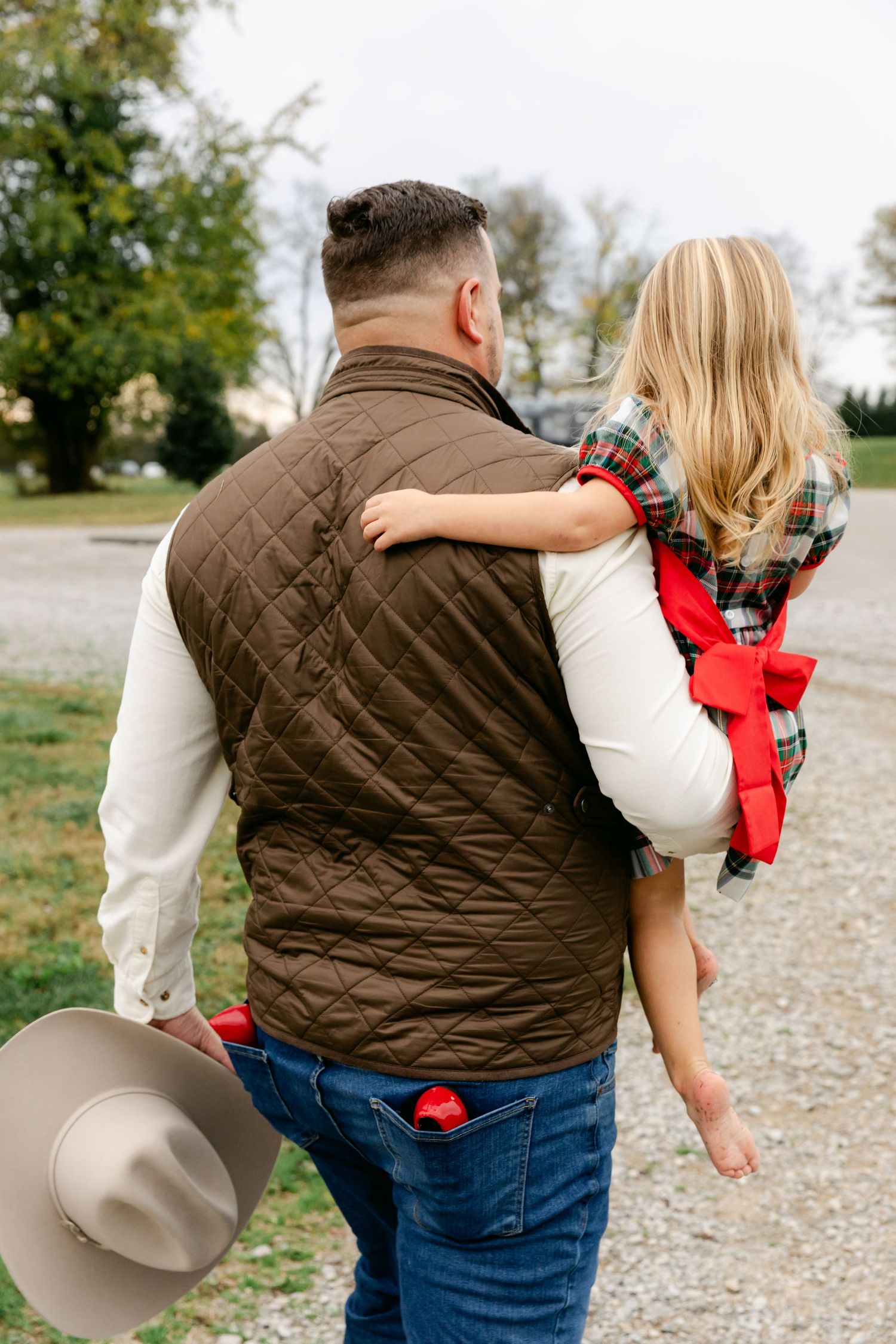Man in brown quilted vest carries young girl in plaid dress with red bow, holding cowboy hat outdoors.