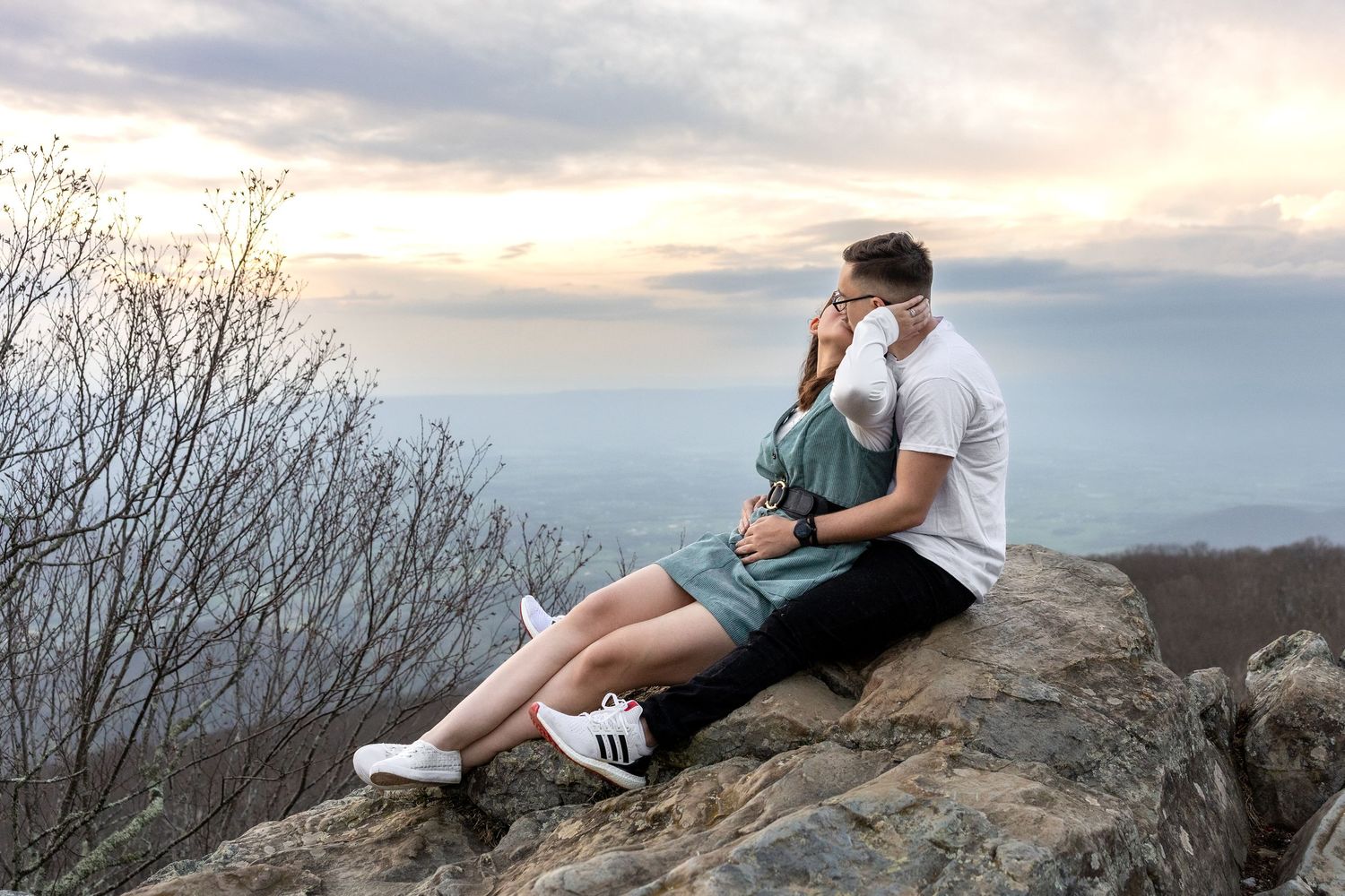 A couple sits together on a rocky mountain overlook during sunset as clouds gather in the dramatic sky.