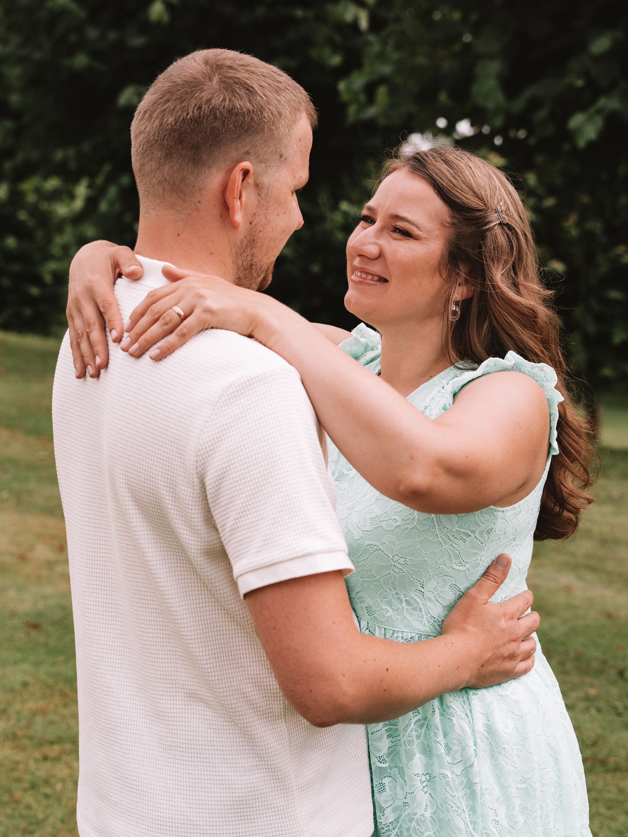 A couple embracing outdoors, smiling at each other, woman in mint dress, man in white shirt, green trees background.