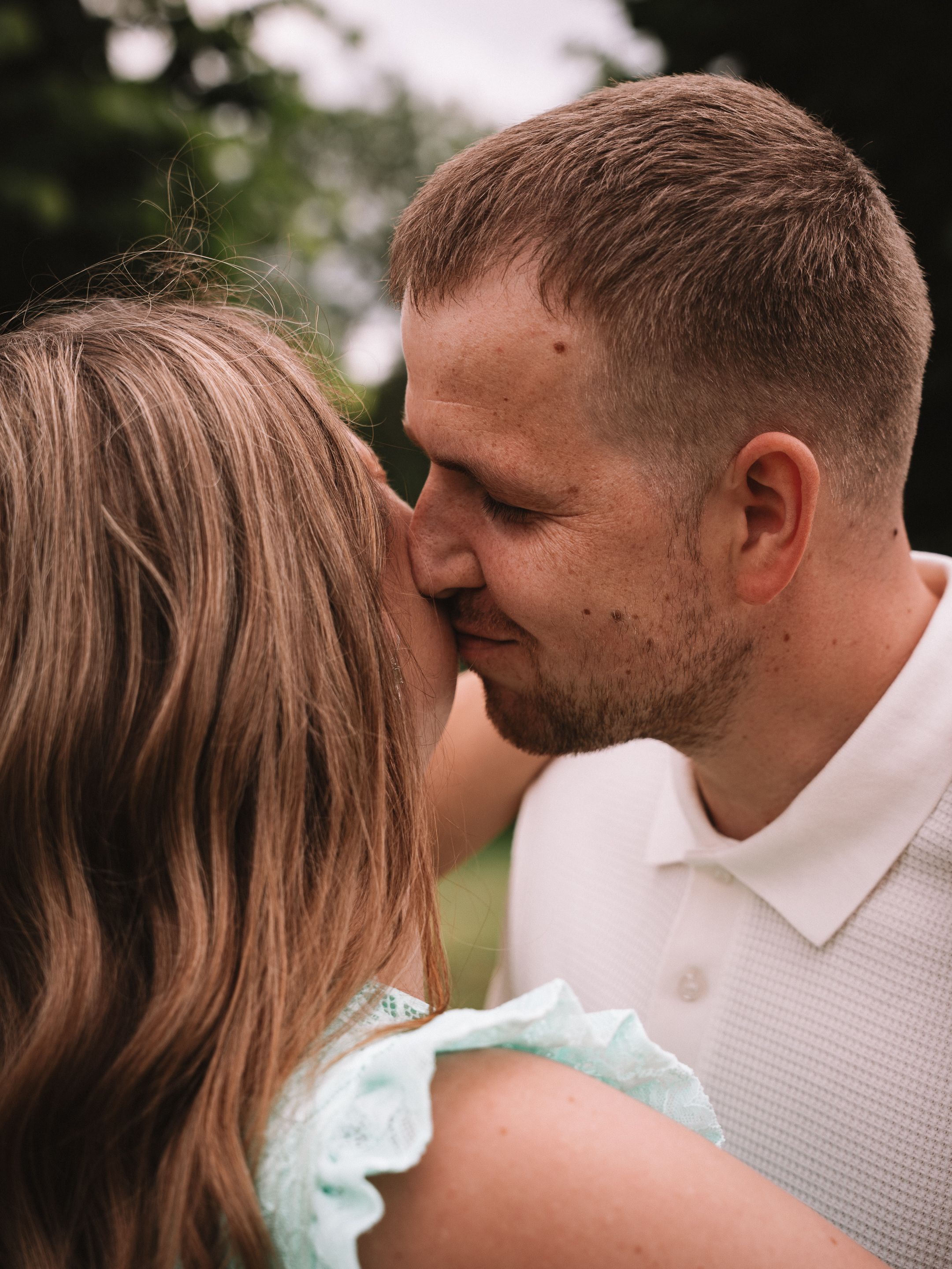 A couple sharing a romantic kiss outdoors, the man in a white polo and woman in a mint ruffle top, surrounded by lush greenery.