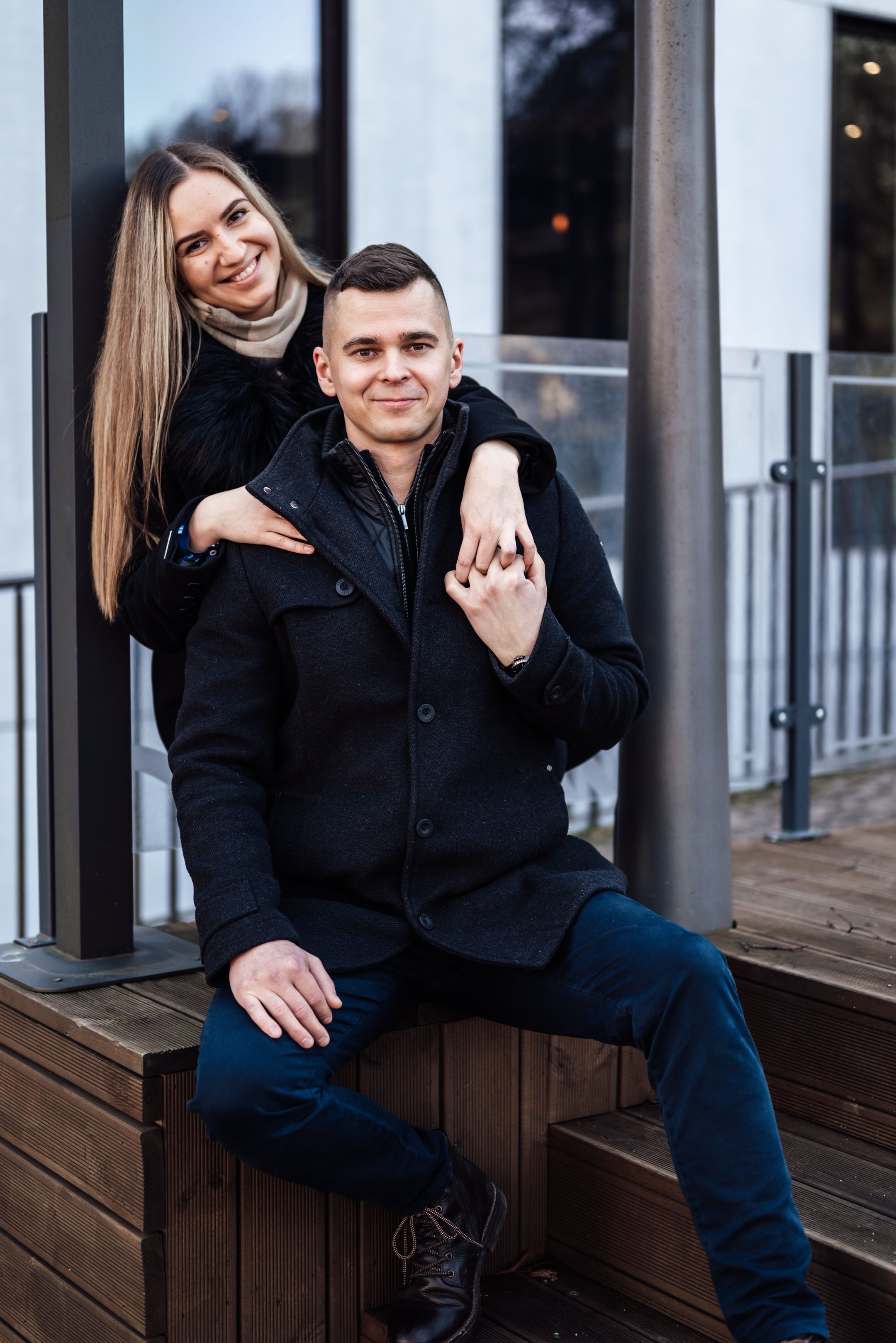 A smiling couple in black coats poses outdoors, woman standing behind seated man on wooden steps.