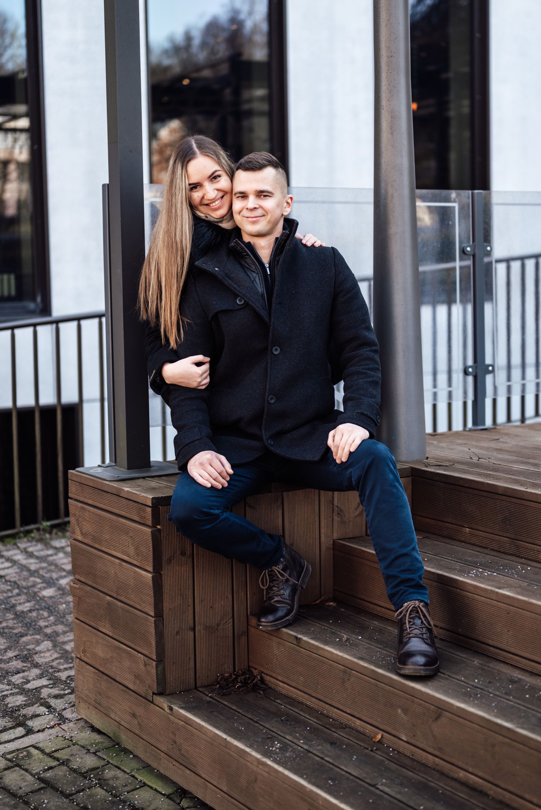 A smiling couple in black coats poses on wooden steps outside a modern building, with the woman embracing the seated man.