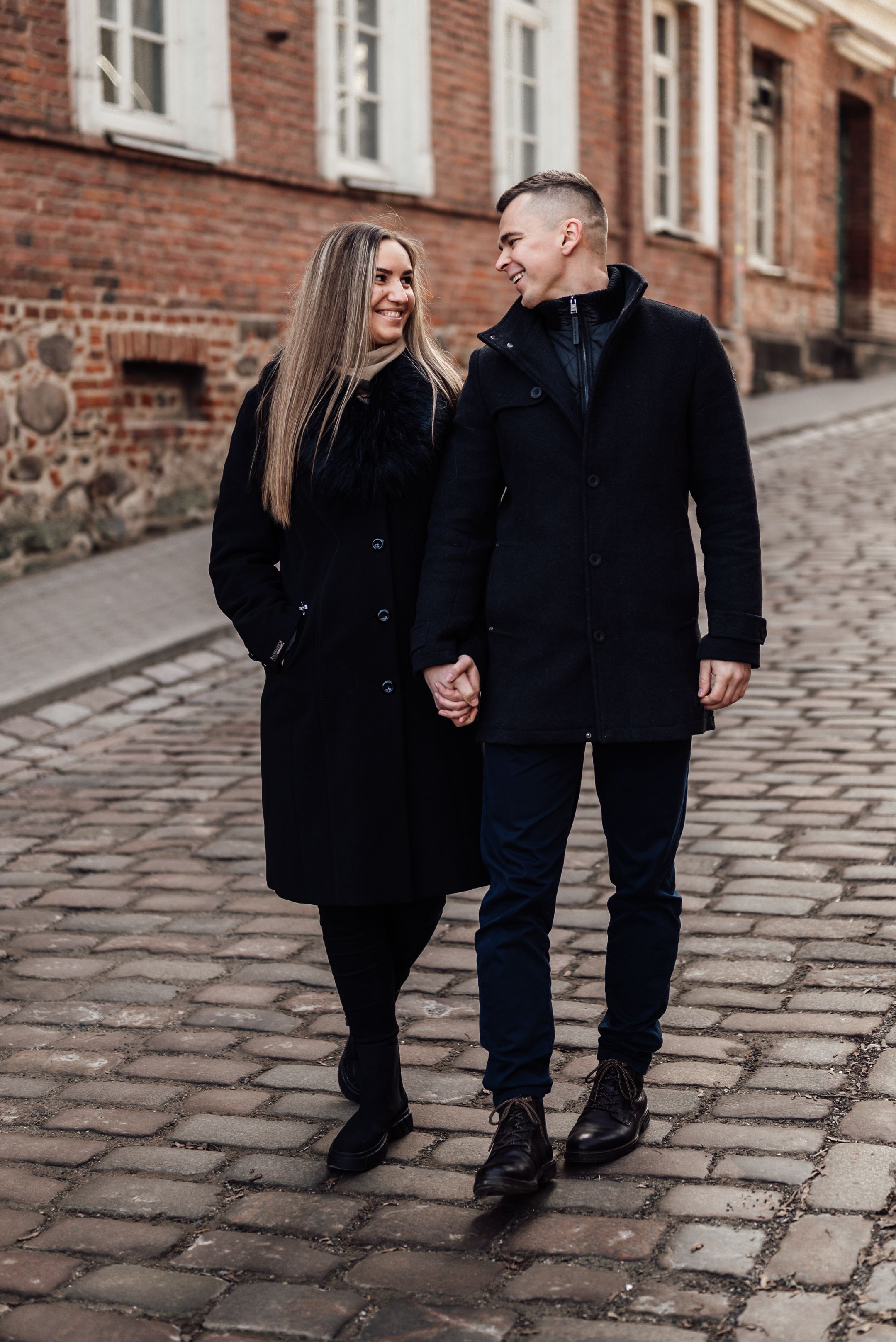 Couple in black coats holding hands, smiling at each other on a cobblestone street near a brick building.