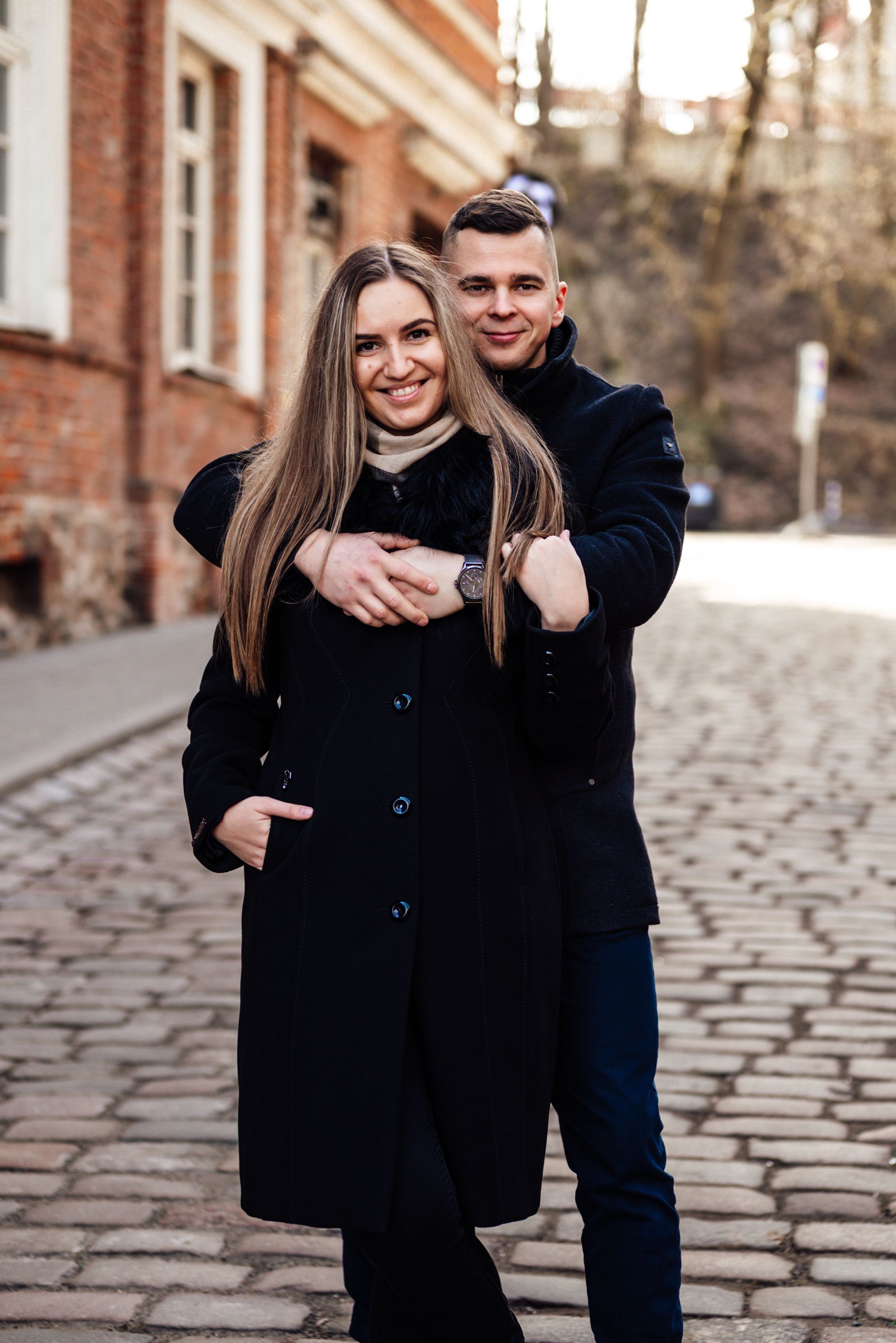 A smiling couple in black coats embracing on a cobblestone street beside a brick building.