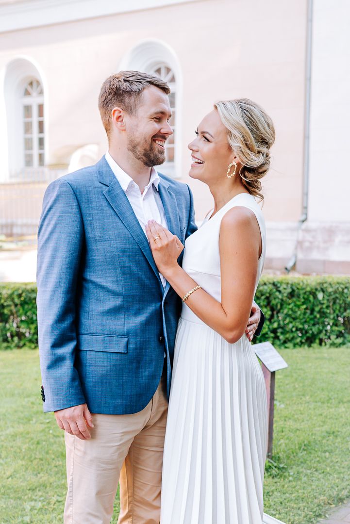 Happy couple in wedding attire laughing together outdoors, man in blue blazer and woman in white pleated dress.