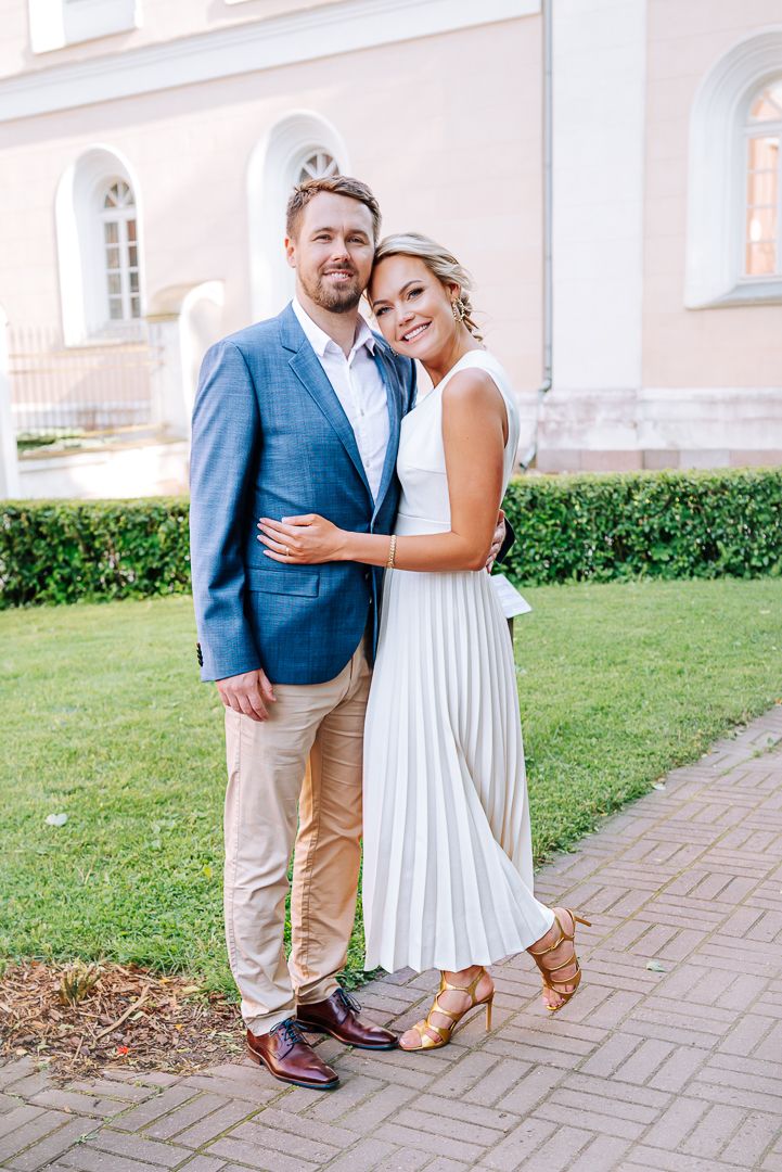 Couple posing outdoors, man in blue blazer and woman in white pleated dress, smiling together near a pink building.