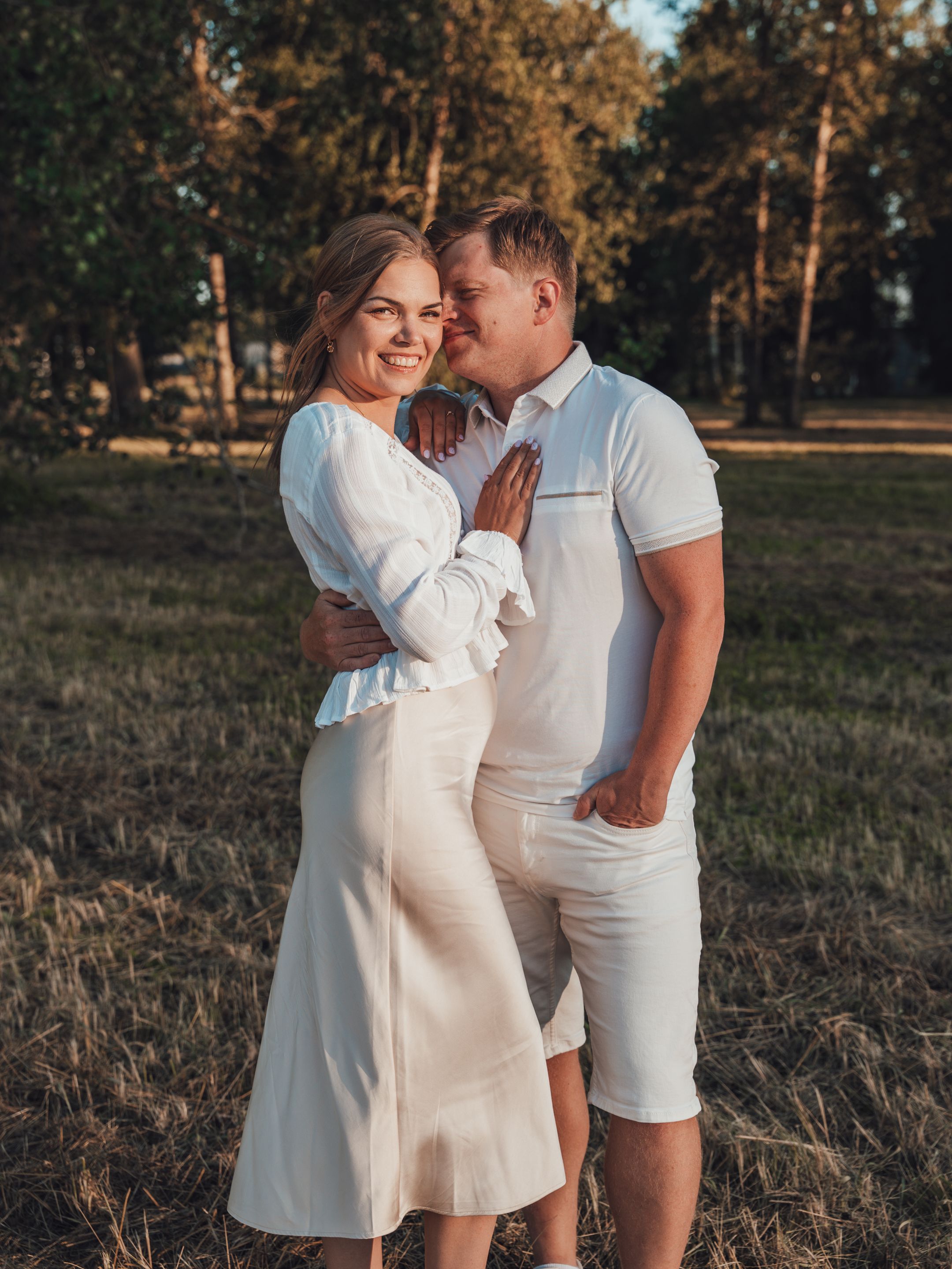 Couple in matching white outfits sharing a romantic moment in a sunlit field with trees in the background.