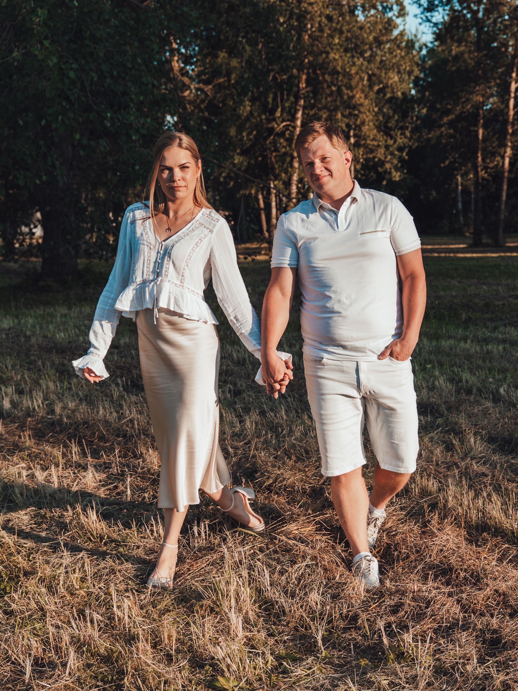 A couple holds hands walking through a sunlit forest clearing, dressed in matching white outfits on a warm day.