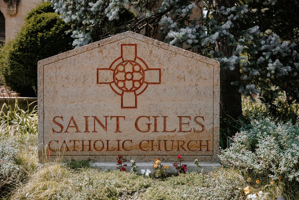 Stone sign for Saint Giles Catholic Church with decorative cross, surrounded by lush greenery and small flowers.