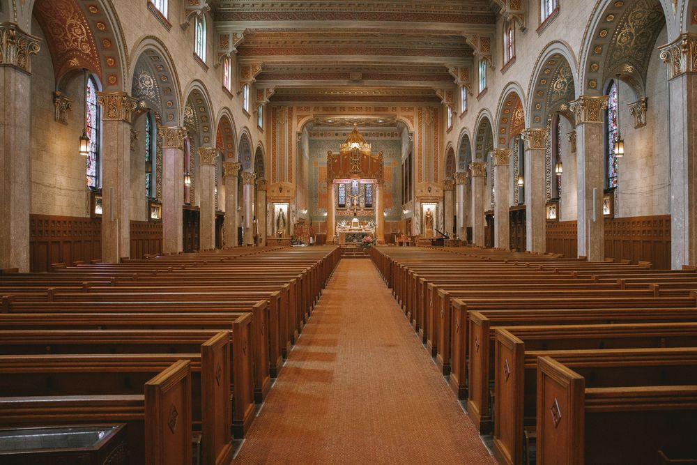 Grand cathedral interior with rows of wooden pews leading to ornate altar, featuring arched ceilings and stained glass windows.