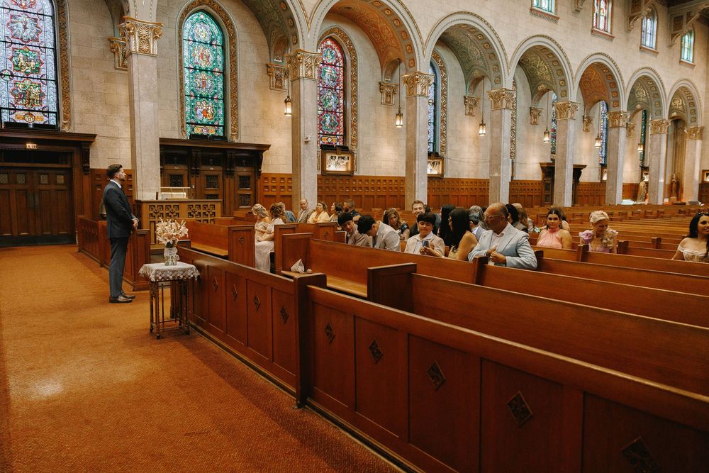 Guests seated in wooden church pews during a wedding ceremony in a grand cathedral with stained glass windows.