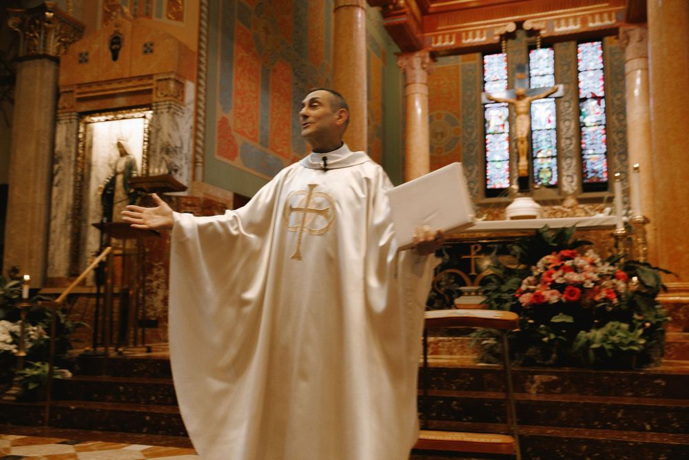 Catholic priest in white vestments with gold cross gestures during Mass at ornate church altar with floral arrangements.